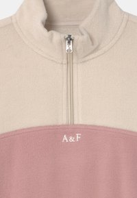 Fleece quarter-zip trui in lichtcrème en zachtroze, met een hoge kraag, zichtbare naden en geborduurd "A&F" logo op de borst.