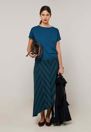 Femme aux cheveux foncés portant un haut bleu et une jupe asymétrique à rayures, tenant une veste foncée et un sac à main marron, debout devant un fond uni.