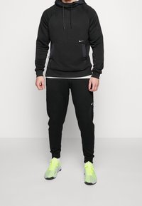 Czarny sportowy hoodie z teksturowanymi panelami i przednią kieszenią, w zestawie z dopasowanymi czarnymi dresami. Jaskrawozielone sneakersy dopełniają stylizację.