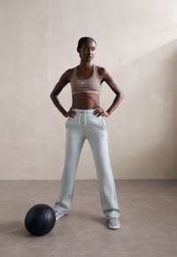 Brūnā sporta krūšturis un gaiši zili treniņbikses, stāv pārliecināti ar rokām uz gurniem blakus melnā medicīnas bumbai uz neitrāla fona.