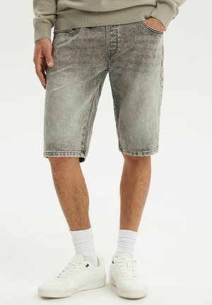 Mężczyzna noszący wyblakłe szare szorty jeansowe, białe skarpetki za kostkę i białe trampki, stojący z jedną ręką w kieszeni.