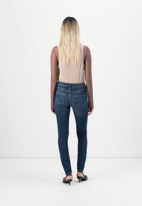 Slanke blauwe jeans met een hoge taille en vier zakken, gecombineerd met een lichtbruine lange mouwtop, gedragen met zwarte haksandalen.