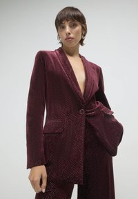 Giacca in velluto bordeaux con scollatura a V profonda, tasca frontale e dettaglio decorativo della cravatta. Presenta delicati motivi testurizzati e chiusura a bottone.