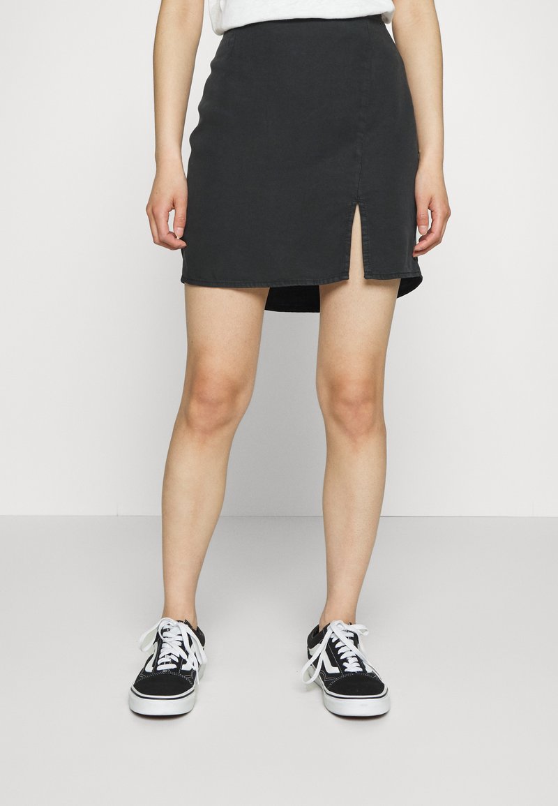 Saia mini de ganga preta com uma racha frontal, apresentando uma bainha limpa e um corte slim. Combinada com ténis pretos e atacadores brancos.