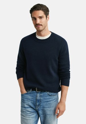 Marineblauer Pullover mit Rundhalsausschnitt und gerippten Bündchen, aus glattem Strickmaterial. Kombiniert mit hellblauen Jeans, lässig getragen.