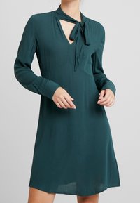 Vestido teal hasta la rodilla con detalle de cuello atado, mangas largas y escote en V. Tela suave y lisa con una silueta fluida.
