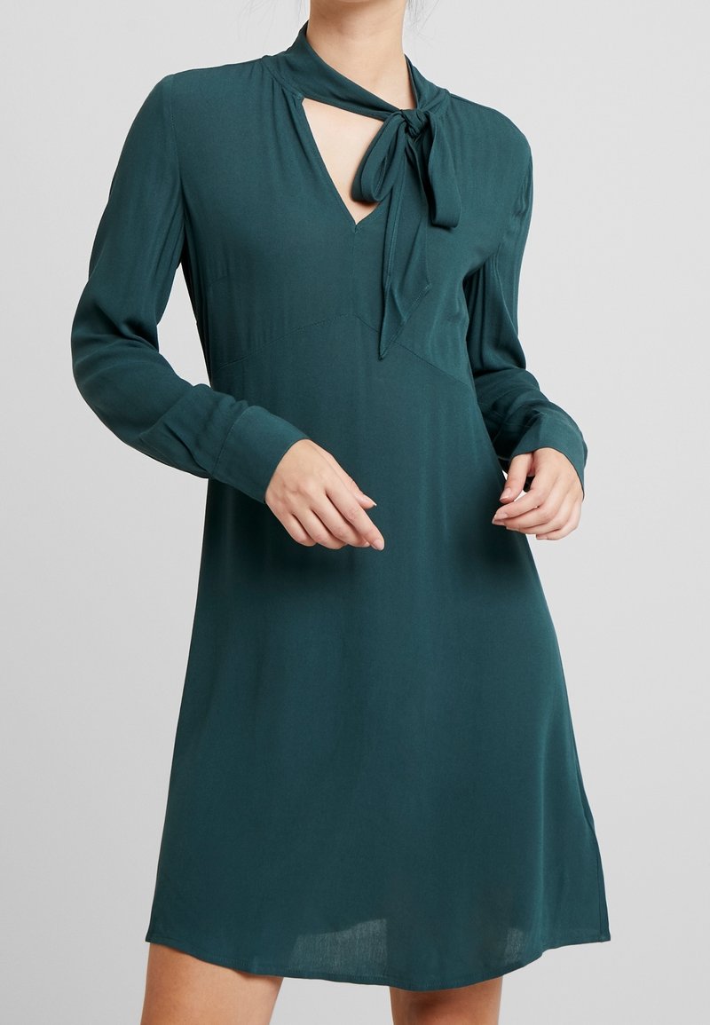 Türkisfarbenes, knielanges Kleid mit einem Bindeband am Hals, langen Ärmeln und einem V-Ausschnitt. Weicher, glatter Stoff mit einer fließenden Silhouette.
