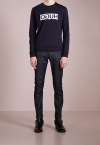 Marineblaues Sweatshirt mit weißem "HUGO"-Logo, kombiniert mit dunklen Jeans und schwarzen Stiefeletten. Glatte Stoffoberfläche, gerippte Bündchen und Saum.