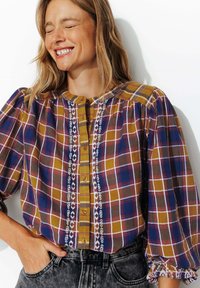 Blouse à carreaux présente des tons jaunes, marron et bleus. Boutonnage sur le devant avec des détails brodés le long de la patte de boutonnage et des manches bouffantes.