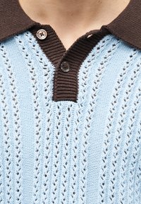Ανοιχτό μπλε πλεκτό πουλόβερ με καφέ ριμπ γιακά και δύο καφέ κουμπιά, φορεμένο στην περιοχή του άνω στήθους και του λαιμού ενός ατόμου.