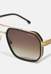 Ochelari de soare cu ramă metalică aurie, accent din cauciuc negru, lentile gradient maro și inscripția 'CARRERA UV PROTECT' gravată pe lentilă.