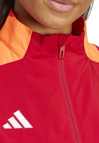 Veste de sport rouge avec col orange, dotée d'une fermeture éclair à l'avant et d'un logo brodé blanc. Texture lisse et design moderne.