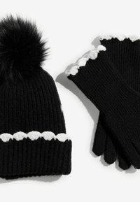 Melnā adīta cepure ar pūkainu pom-pomu un baltu viļņotu maliņu, saskaņota ar melniem cimdiem, kas arī ir ar viļņotu baltu apdari.