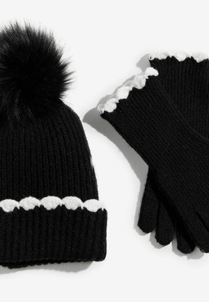 Melnā adīta cepure ar pūkainu pom-pomu un baltu viļņotu maliņu, saska�ņota ar melniem cimdiem, kas arī ir ar viļņotu baltu apdari.