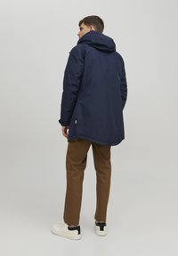Giacca navy con cappuccio, silhouette lunga e polsini elasticizzati. Indossata sopra pantaloni marroni e sneakers bianche con dettagli neri.