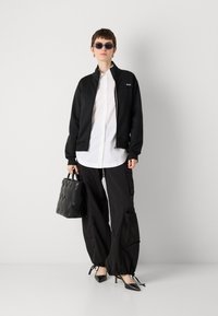 Black zip-up jacket with high collar, white button-up shirt, black cargo pants with zippers, pointed black heels, and a textured black handbag.