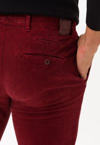 Pantalon en velours côtelé rouge avec un design texturé et côtelé, doté de poches arrière et d'une étiquette en cuir marron. Coupe slim avec une apparence taillée.