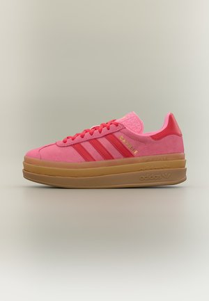 Baskets Adidas Gazelle roses avec des bandes rouges, inscription Gazelle dorée, semelle épaisse couleur beige et languette rose texturée sur fond neutre.