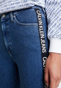 Vaqueros de denim en un tono azul oscuro; cuentan con una tira lateral contrastante en negro con "CALVIN KLEIN JEANS" en texto blanco; diseño clásico de cinco bolsillos.