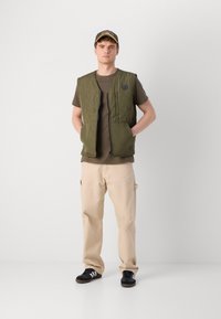 Olive green quilted vest with two front pockets, worn over a brown t-shirt and beige pants, paired with black sneakers and a cap.