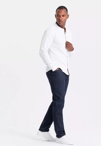 Camisa blanca de manga larga con cuello y ribete en contraste, combinada con unos vaqueros azul oscuro y zapatillas blancas, exhibida en un modelo de pie.