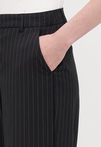 Mustat pinstripet housut, joissa on litteä etuosa, sivutaskut ja sileä kangas. Kankaassa on ohuita valkoisia viivoja, jotka kulkevat pystysuoraan.