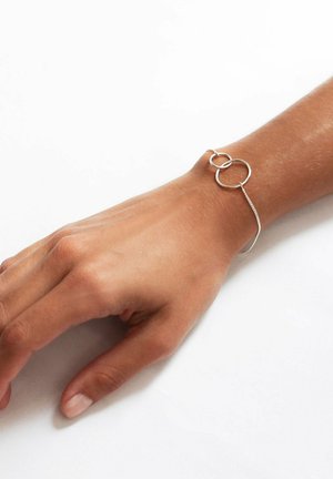 Pulsera de plata con dos círculos entrelazados; presenta una cadena fina y un acabado suave. Lleva puesta en una mano de tono de piel claro.