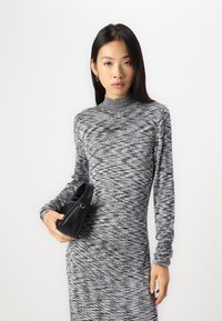 Pilkai ir juodai raštuotas ilgos rankovės suknelė su aukštu apykakle, ant kurios matyti logotipas „Karl Lagerfeld Jeans“. Modelis laiko mažą juodą rankinę.