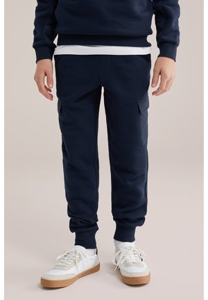 Személy tengerészkék futónadrágot visel oldalzsebekkel, fehér zoknit és fehér, gumitalpú sportcipőt viselve, egyszerű háttér előtt áll.