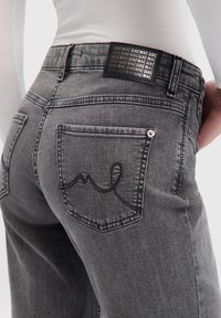 Jean en denim gris avec un détail de poche arrière cachée et une étiquette de marque noire. Présente des coutures ton sur ton et une texture légèrement délavée.