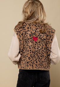 Bruine fleecevest met zwarte luipaardvlekken, voorzien van een rode hart en geborduurde tekst op de achterkant, gecombineerd met een beige langemouwenshirt.