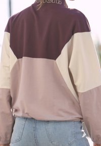 Veste de sport bicolore en violet, crème et rose clair, confectionnée dans un tissu lisse, avec un col montant et des détails de coutures visibles.