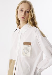 Weißes Hemd mit Knöpfen, das am unteren Ende über ein beiges Panel, eine Tasche mit Etikett und einen schwarzen Akzent am Kragen verfügt. Glatte Textur.