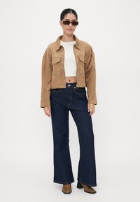Sandfarget semsket jakke med cropped design og frontlommer, kombinert med mørkeblå slengjeans. Modellen har på seg klare solbriller og står mot en hvit bakgrunn.