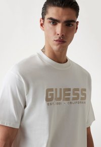 Weißes Baumwoll-T-Shirt mit großem beigen "GUESS"-Logo und dem Text "EST.1981 - CALIFORNIA" auf der Vorderseite, rundem Ausschnitt und kurzen Ärmeln.