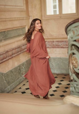 Rochie de culoare ruginiu, fluidă, cu mâneci largi și tăietură la spate. Materialul neted și ușor se așează elegant în timp ce modelul urcă scările.