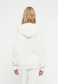 Bijela rebrasta hoodie s velikom, mekanom, zaobljenom kapuljačom. Ima duge rukave i opušteni kroj. Tekstura izgleda udobno i toplo.