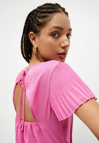 Blusa rosa con maniche corte a pieghe, schiena aperta fissata con chiusura a fiocco e una texture di tessuto liscia. Orecchini a cerchio dorati aggiungono un tocco di dettaglio.