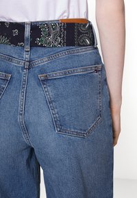 Džínové kalhoty s tmavým páskem s potiskem bandany a hnědou koženou štítkem. Obsahuje klasické zadní kapsy a podrobný design švu.