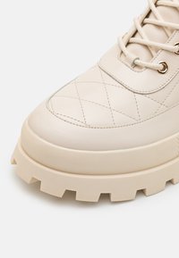 Botte beige à lacets avec dessus en cuir matelassé, œillets dorés et semelle en caoutchouc épaisse et robuste avec des crampons profonds.