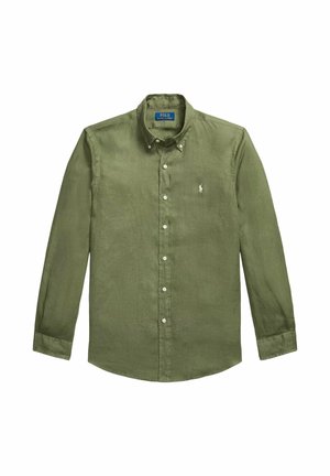 Homme portant une chemise et un pull vert olive, pantalon blanc avec cordon, baskets blanches, tenant un sac cabas en cuir marron, regardant vers le bas, main dans la poche.