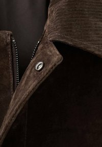 Ovratnik smeđe jakne od antilopa s teksturiranim tkanjem, detaljem patent zatvarača na lijevoj strani i ukrasom od kopče na pritisak. Vidljiva je glatka unutrašnja podstava.