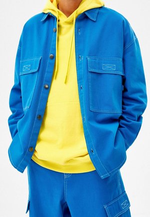 Person trägt eine leuchtend blaue Jacke und Hose mit großen Taschen über einem gelben Kapuzenpullover mit Kordelzügen.