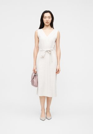 Donna con capelli lunghi e scuri che indossa un abito midi bianco senza maniche con cintura in vita, che tiene una piccola borsa rosa chiaro, in piedi davanti a uno sfondo semplice.