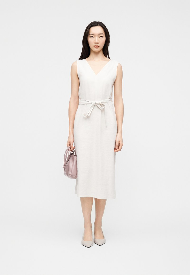 Γυναίκα με μακριά σκούρα μαλλιά που φορά λευκό μίντι φόρεμα χωρίς μανίκια με ζώνη στη μέση, κρατώντας μικρό ανοιχτό ροζ τσαντάκι, στέκεται μπροστά σε απλό φόντο.