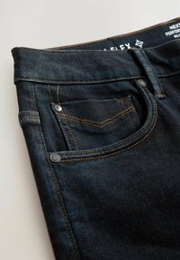 Nærbillede af en lomme på mørkeblå denimjeans med orange syninger, metalnitter, bæltestrop og delvis tekst på linningen indeni.