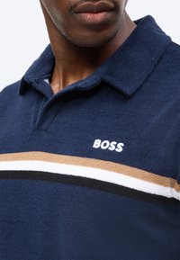 Κοντινό πλάνο ενός ατόμου που φοράει μπλε σκούρο πουκάμισο με γιακά, με μπεζ, λευκές και μαύρες ρίγες και τη λέξη "BOSS" κεντημένη στο στήθος.