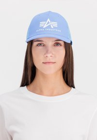 Kék baseball sapka fehér logóval, amelyen az "ALPHA INDUSTRIES" felirat látható, sima fehér pólóval párosítva. A dizájn struktúrált, hálós háttal rendelkezik.