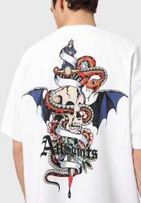 Άσπρο βαμβακερό μπλουζάκι με μεγάλο γραφικό ενός κρανίου με φίδια, λουλούδια και κορδέλες σε ζωντανά χρώματα και περίπλοκες λεπτομέρειες.