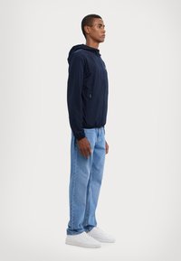Marineblå hættejakke med en lynlåslomme, elastiske manchetter og et let design, parret med lyseblå jeans og hvide sneakers.